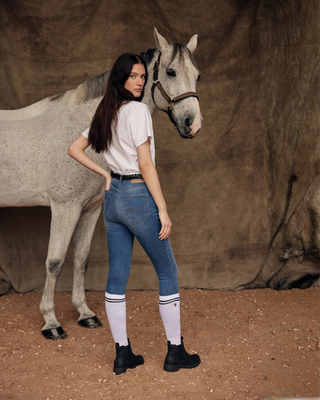 Sfeervolle lifestylefoto van een grijs paard en model met een wit T-shirt, OG denim rijbroek en witte sokken.