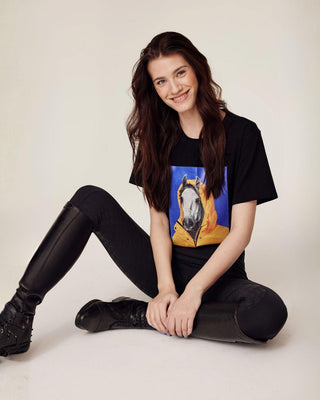 Ontspannen zittend model dat de zwarte rijbroek draagt, gecombineerd met een zwart t-shirt met paardenprint.