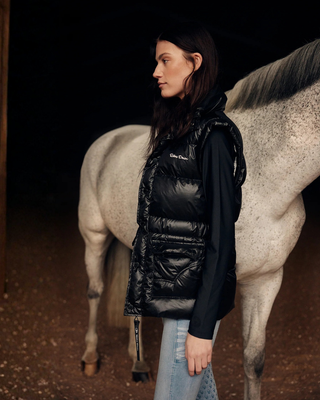 Lifestyle-opname van het model van opzij met een grijs paard, gekleed in de zwarte pufferbodywarmer en zwarte rijtop.