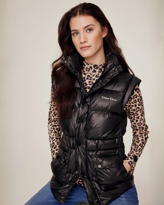 Zittend model dat het zwarte halfglanzende pufferbodywarmer draagt, dichtgeritst om de functionele tweewegritssluiting te tonen, gestyled met een leo top en denim rijbroek.