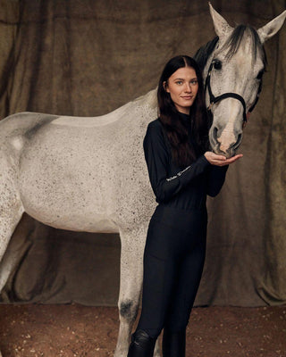 Sfeerbeeld uit de paardensport van een model in de volledig zwarte rijset dat knuffelt met een grijs paard.