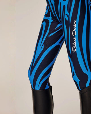 Gedetailleerde opname van het linkerbeen met het witte "Riders Denim"-logo op de blauwe zebraprint.