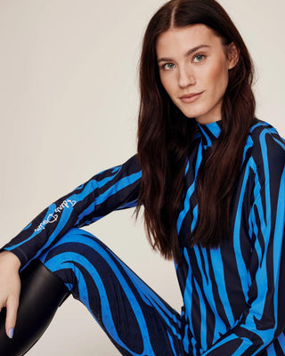 Zittend model poseert in de complete blauwe zebra-rijset en toont het performance-materiaal met vierwegstretch.