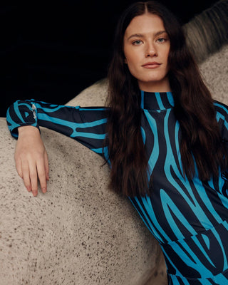 Ontspannen lifestylebeeld van een model dat tegen een grijs paard leunt en de blauwe zebra-print paardensporttop draagt.