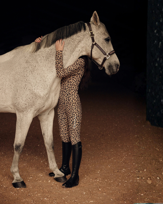 Sfeervolle lifestylefoto van een model dat de volledige luipaard rijset draagt en een grijs paard omhelst.
