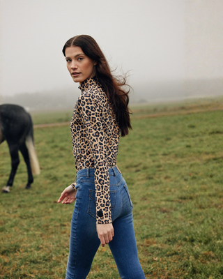 Lifestylefoto van een model dat door een veld loopt in de Leo rijtop en OG denim rijbroek, terwijl ze achterom kijkt.