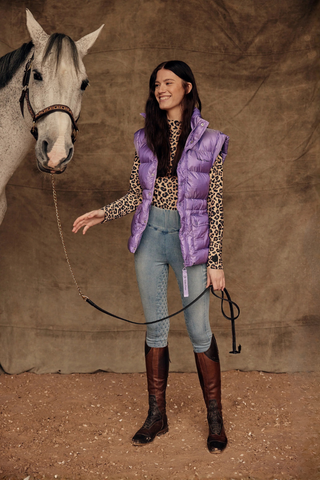 Model in lichtblauwe jeansrijbroek, leo top en lila puffer bodywarmer, terwijl ze een grijs paard aan het halstertouw vasthoudt.