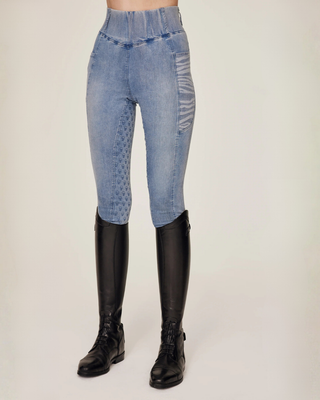 Schuin vooraanzicht van de lichtblauwe denimrijbroek, gestyled met professionele zwarte rijlaarzen.