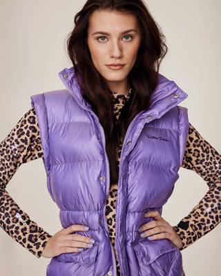 Vooraanzicht van een model dat het semi-glanzende lila riding puffer vest draagt over een riding top met luipaardprint, met de handen in de zij.