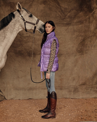 Lifestylefoto van een model in een lila puffer vest, leo top en OG denim rijbroek, staand met een grijs paard.