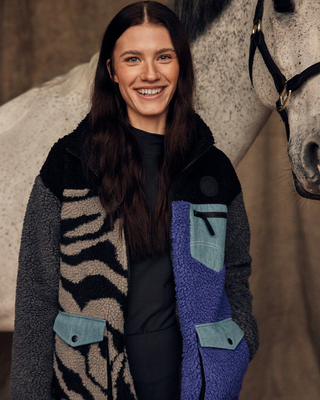 Glimlachend model in een stalomgeving met een open lila en zebra teddyjack, met een grijs paard op de achtergrond.