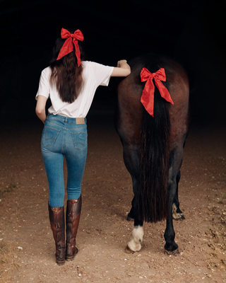 Model van achteren in jeans rijbroek en wit t-shirt met een rode haarstrik, naast een bruin paard met een bijpassende rode strik.
