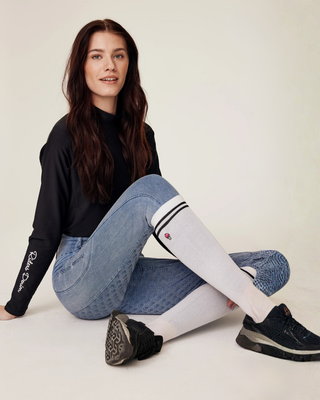 Model poseert in een ontspannen zittende houding, waarbij de complete paardensportoutfit met witte rijsokken wordt benadrukt.