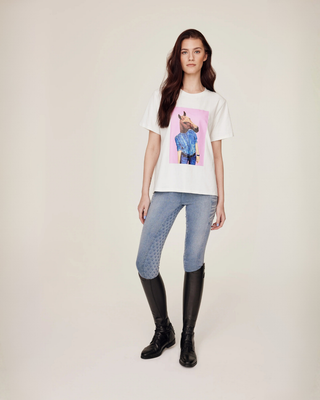 Volledige lichaamsfoto van een model dat het witte T-shirt met paardenprint en lichtblauwe jeansrijbroek toont.