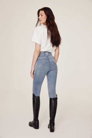 Volledig achteraanzicht van een model in lichtblauwe denimrijbroek en een wit T-shirt met paardenprint, terwijl ze over haar schouder kijkt.